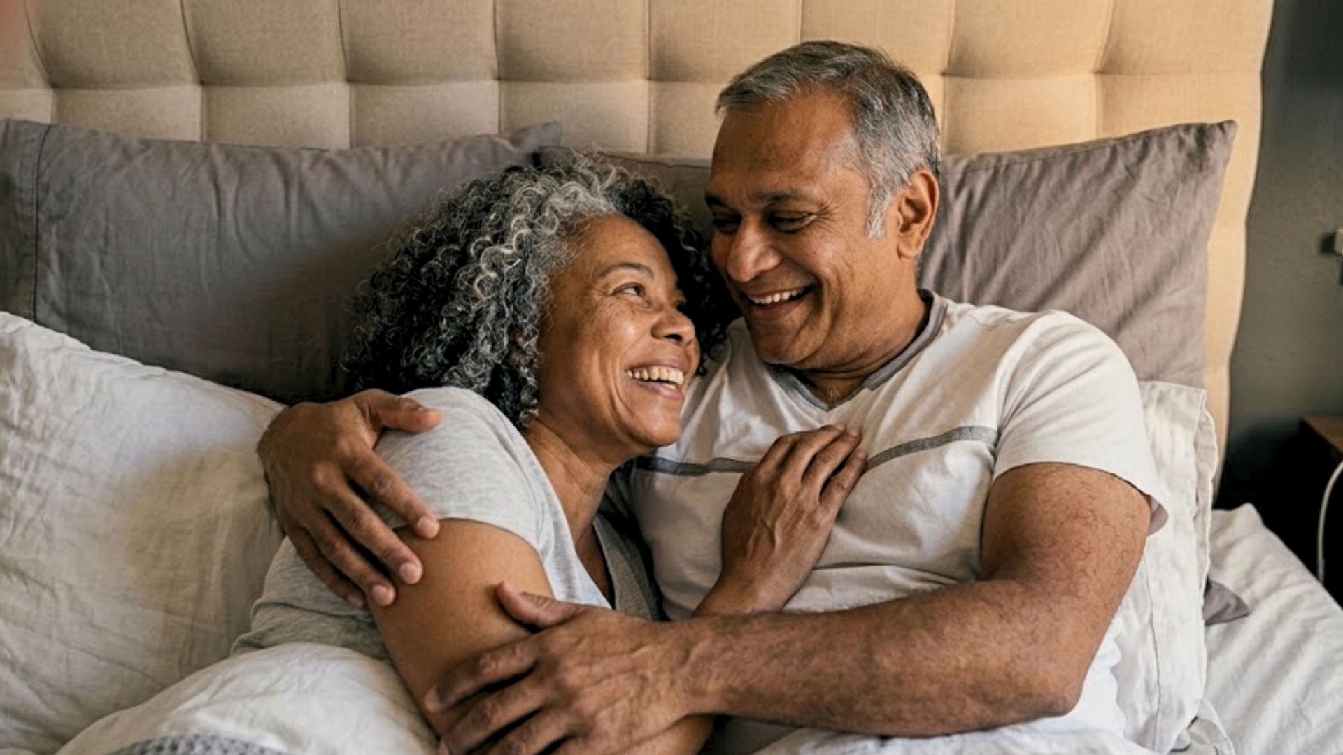 A smiling older couple embraces while lying in bed, looking at each other.
