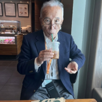 An elderly man in a blue blazer and glasses sips a beverage through a straw in a cafe.