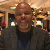 A headshot of a smiling, bald Black man with a gray goatee, wearing a gray blazer.