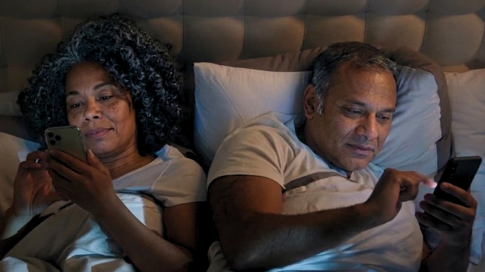 A man and a woman lie in bed next to each other at night, both looking at their smartphones.