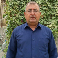 A middle-aged man with glasses and a mustache in a blue button-down shirt, in front of a leafy wall.