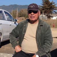 An older man in a cap and sunglasses sits outdoors next to a silver car with hills in the background.
