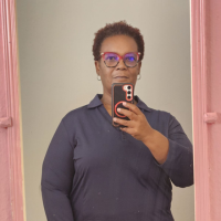 A woman with short, reddish-brown hair and glasses takes a selfie in a mirror with a pink frame.