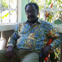 A man with glasses and a mustache, wearing a colorful patterned shirt, sits in a chair.