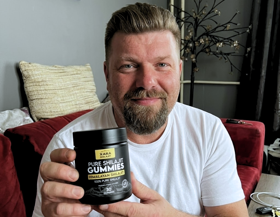 A smiling man in a white t-shirt holds up a black jar of Xara Pure Shilajit Gummies.