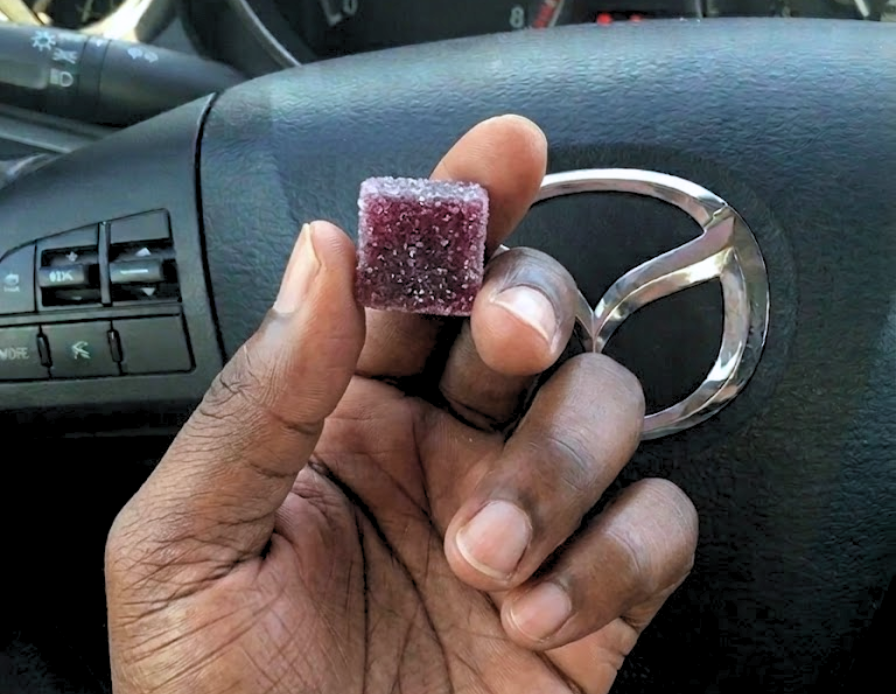 A close-up of a hand holding a purple, sugar-coated gummy in front of a car's steering wheel.