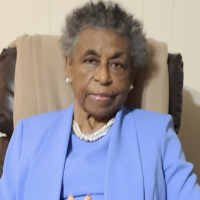 An elderly Black woman with short gray hair, wearing a blue jacket and a pearl necklace, sitting in a chair.