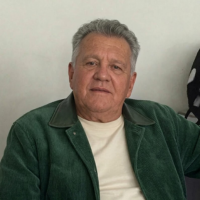 A headshot of a man with graying hair wearing a green jacket over a white t-shirt.