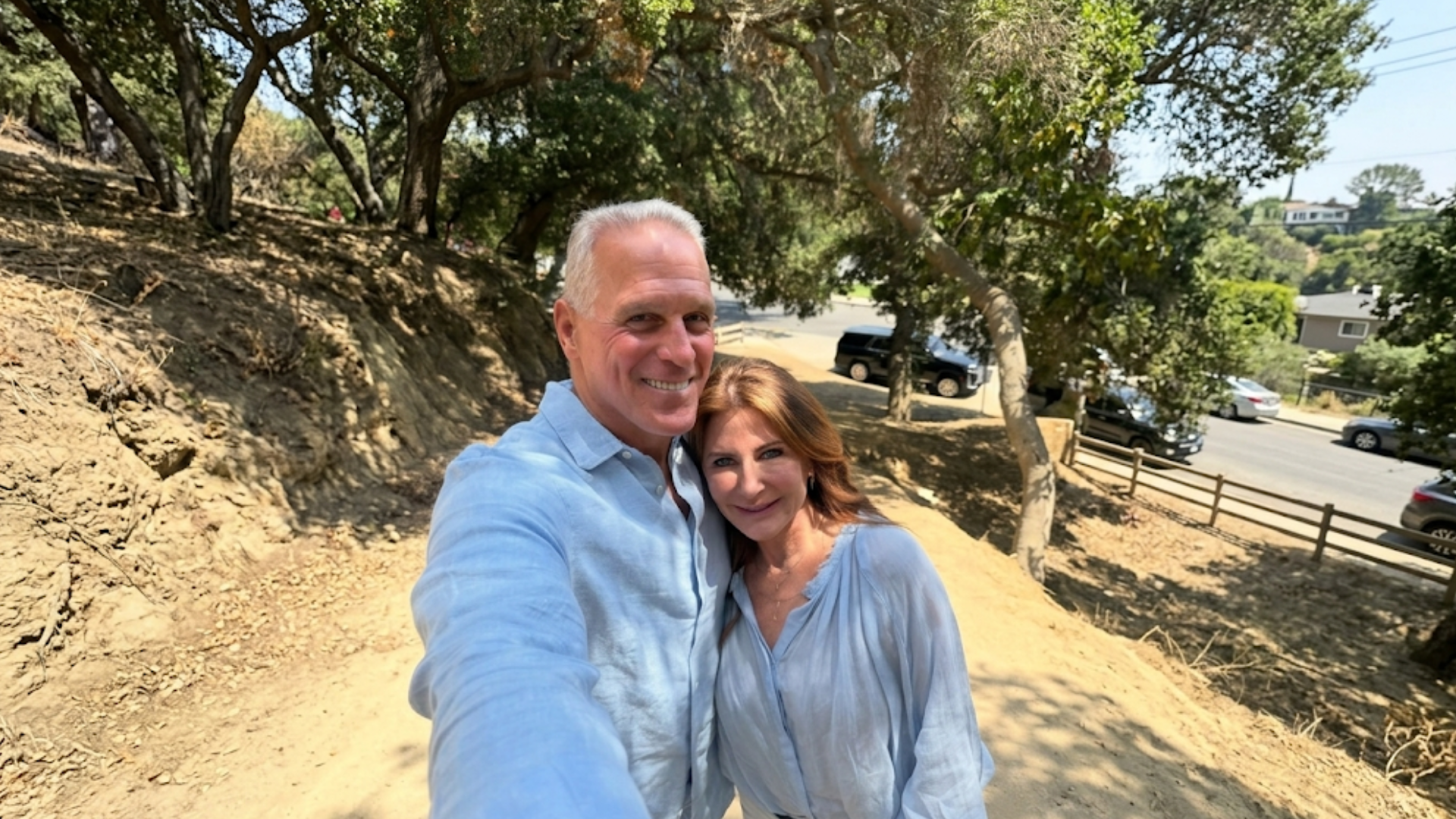 A smiling man and woman take a selfie outdoors on a sunny, wooded hillside trail.