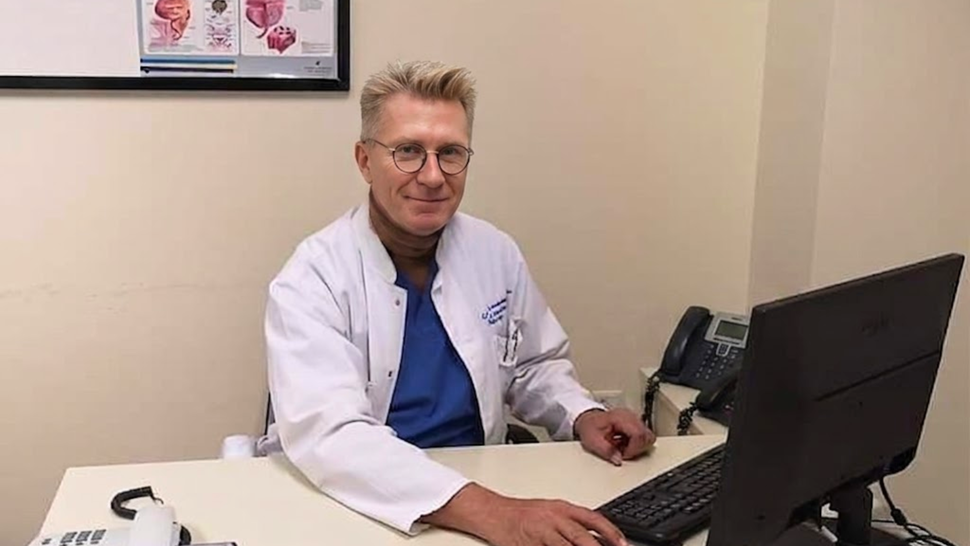 A smiling doctor with glasses and blond hair sits at a desk in a white coat.