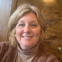 A headshot of a smiling woman with short blonde hair, wearing a brown turtleneck and sweater.