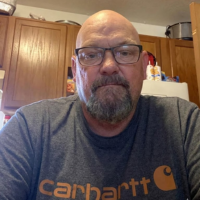 A bald man with a gray goatee and glasses wearing a gray Carhartt t-shirt in a kitchen.