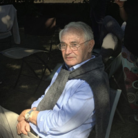 An older man with glasses and a sweater draped over his shoulders sits outdoors in a chair.
