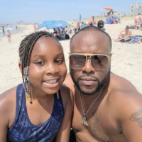 A man and a young girl with braided hair smiling for a selfie on a crowded beach.