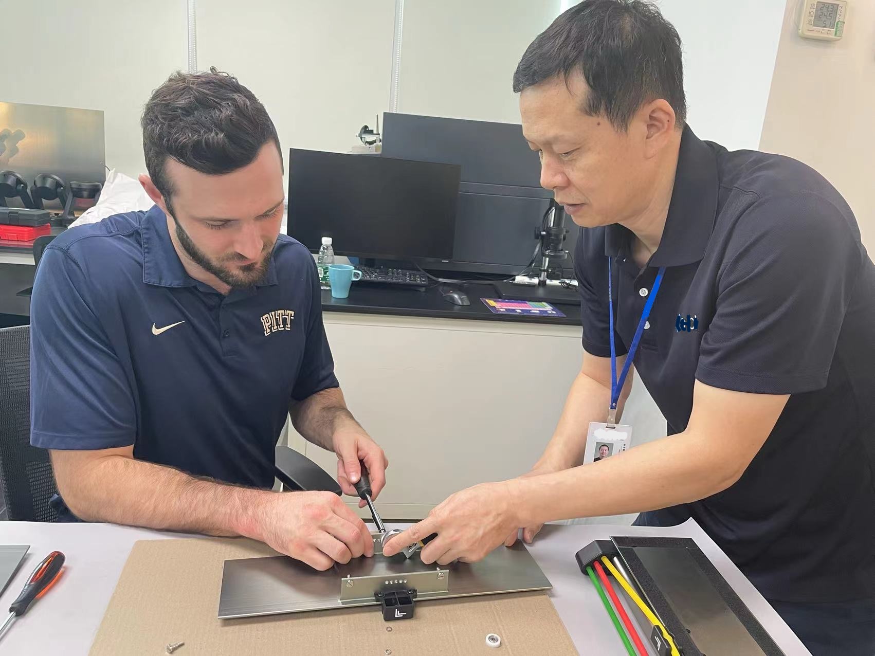 Two people assembling a metal plate with tools on a table.