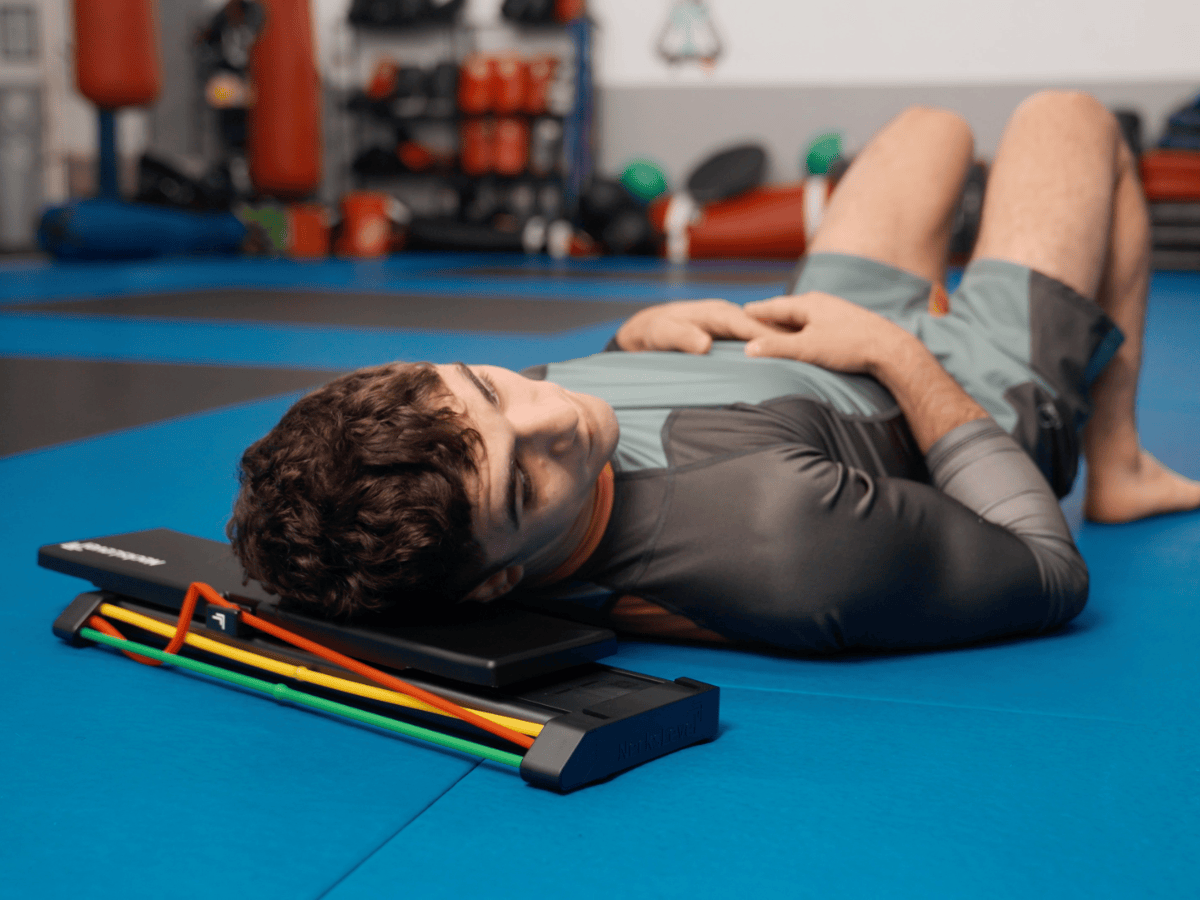 A man lies on his back on a blue mat, using a piece of neck exercise equipment.