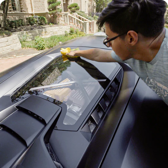 A man wipes the glass engine cover of a matte black sports car with a yellow cloth.