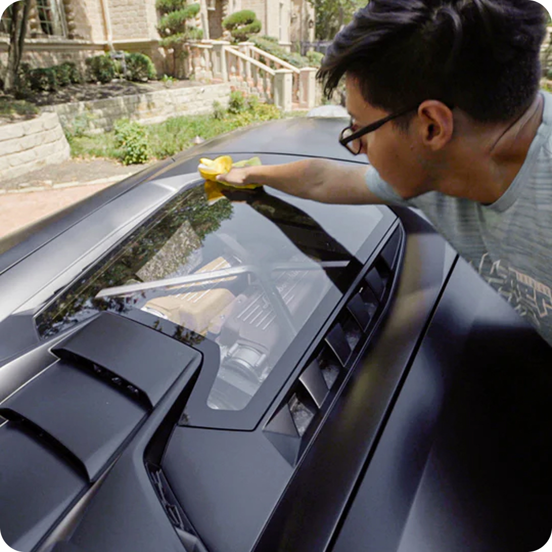 A man with glasses wipes down the rear window of a matte black sports car with a yellow cloth.