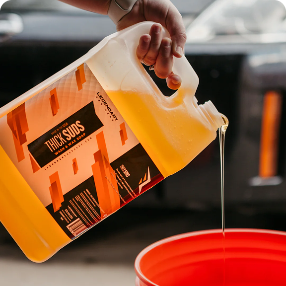 A hand pouring a jug of yellow 'Thick Suds' car wash soap into a red bucket.
