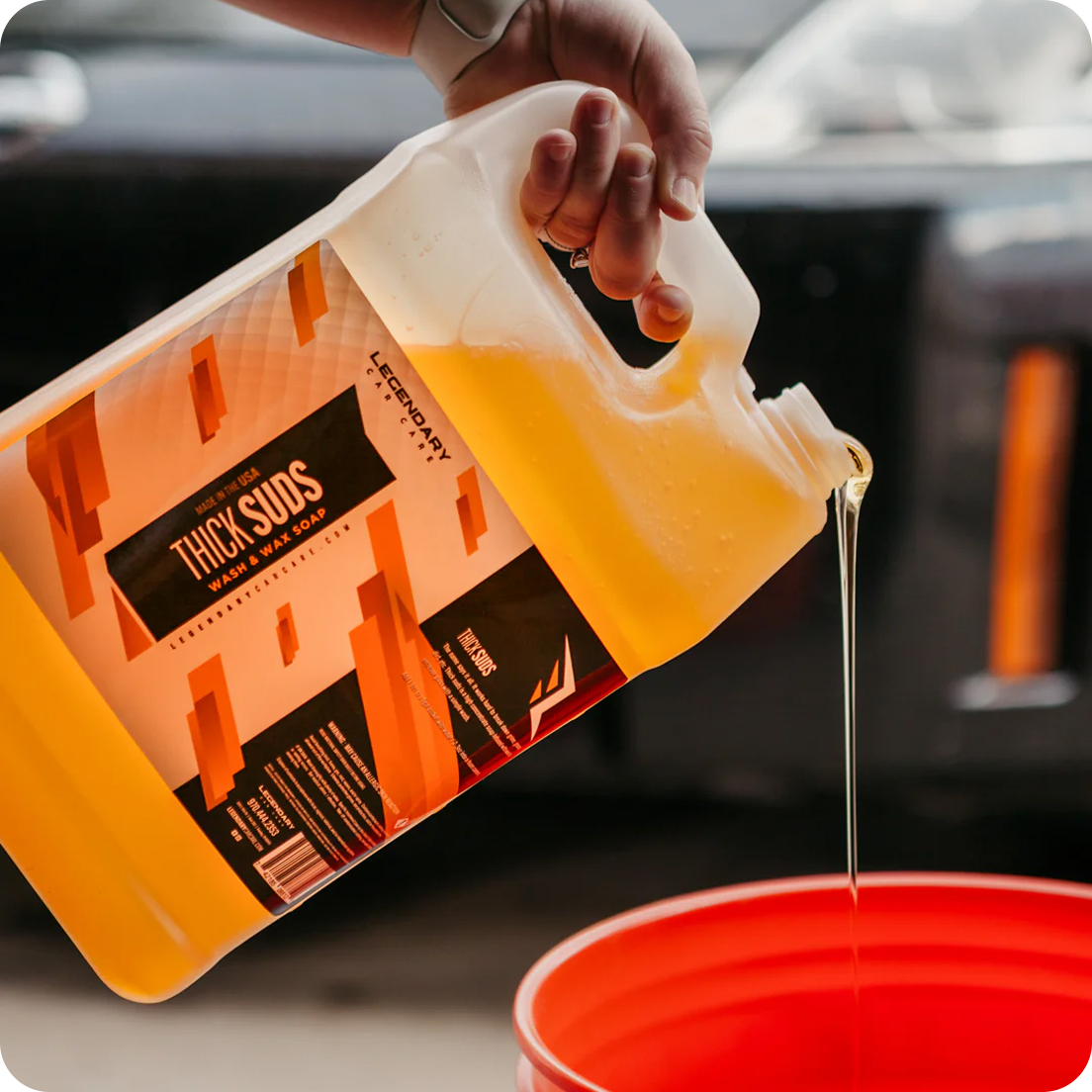 A hand pours yellow Thick Suds wash and wax soap from a large jug into a red bucket.
