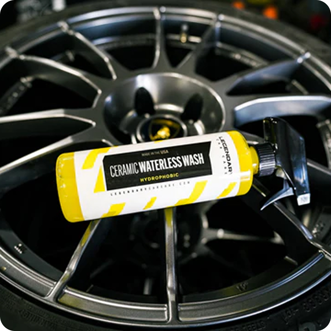 A spray bottle of Ceramic Waterless Wash car cleaner resting on a dark grey multi-spoke car wheel.