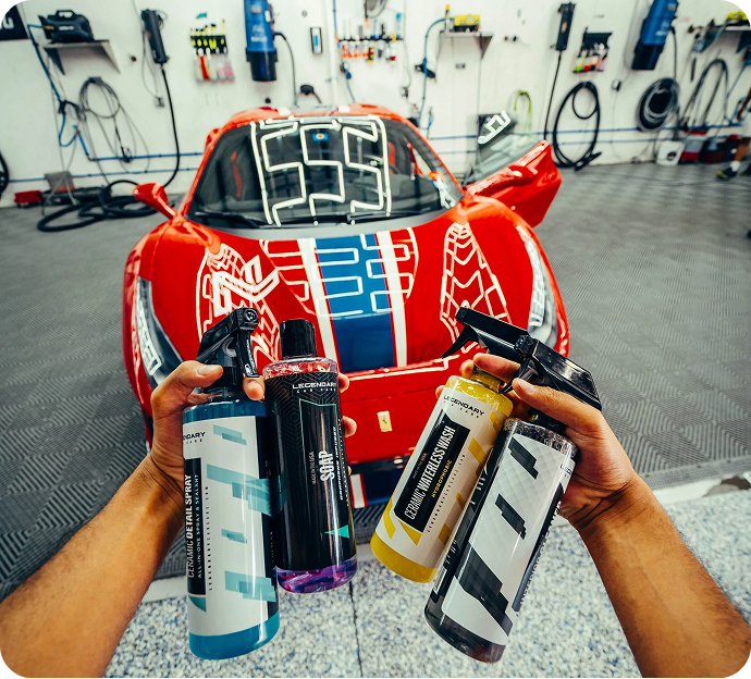A person holds several car cleaning products in front of a red sports car in a garage.