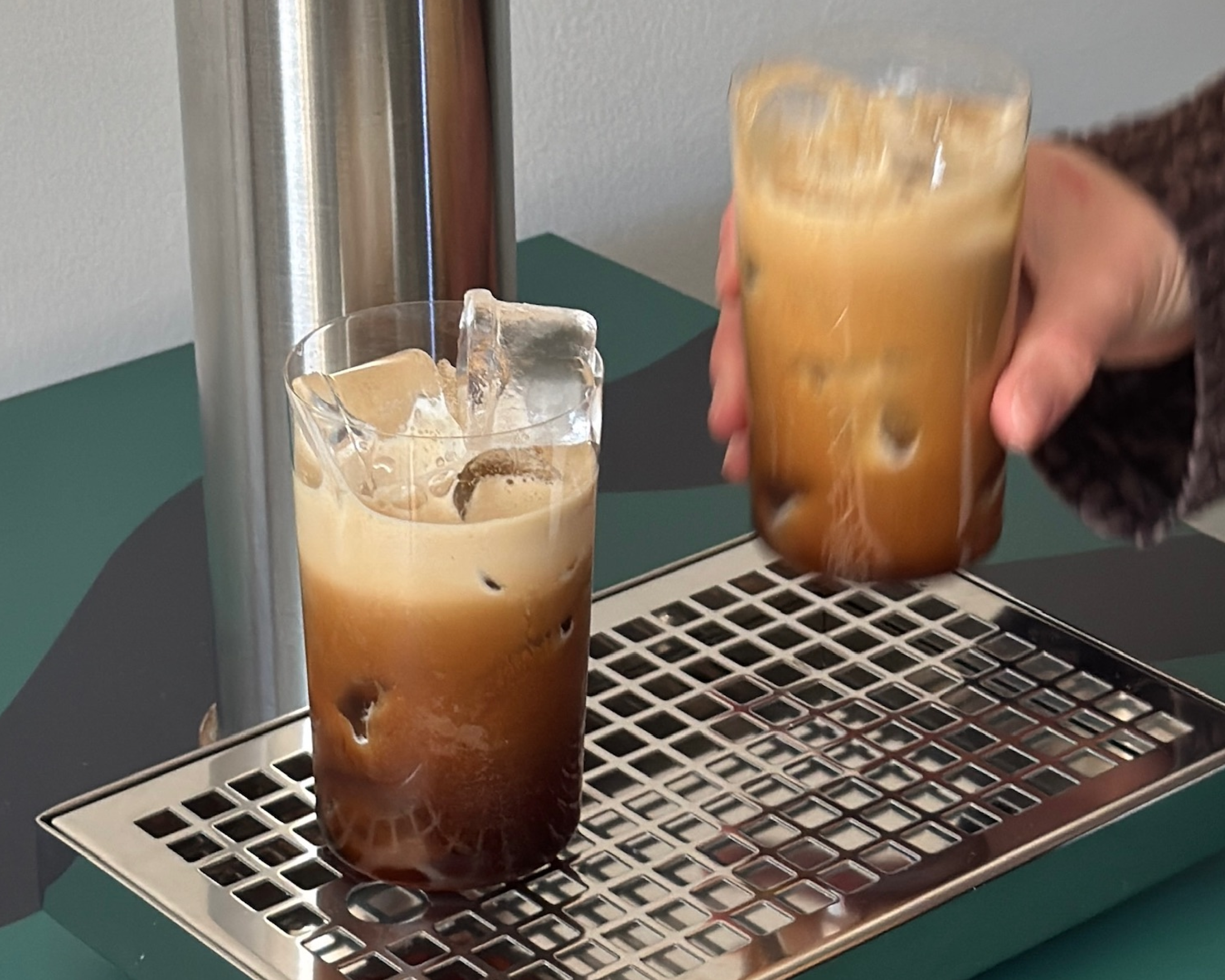 Two glasses of iced coffee on a metal grate with one being held by a hand.