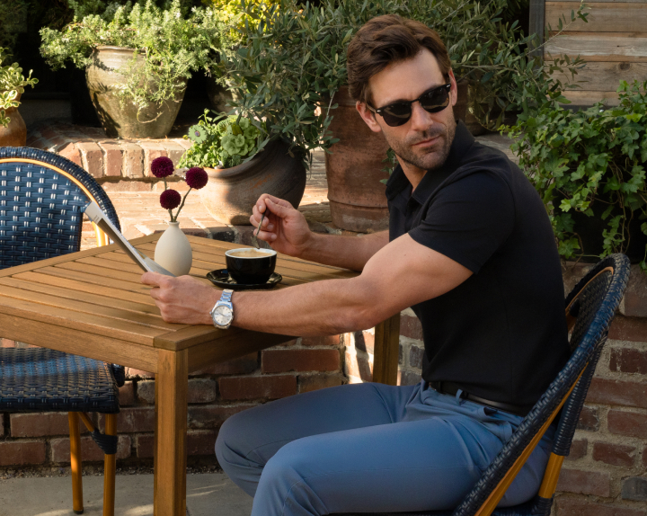 Man in sunglasses sitting at outdoor cafe table with coffee and newspaper.