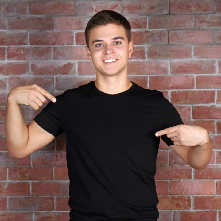 Person in black shirt pointing at themselves against a brick wall background.
