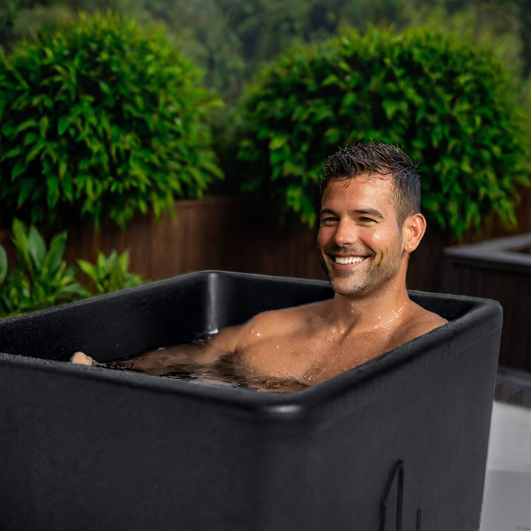 Man smiling while sitting in a black tub outdoors with greenery in the background.