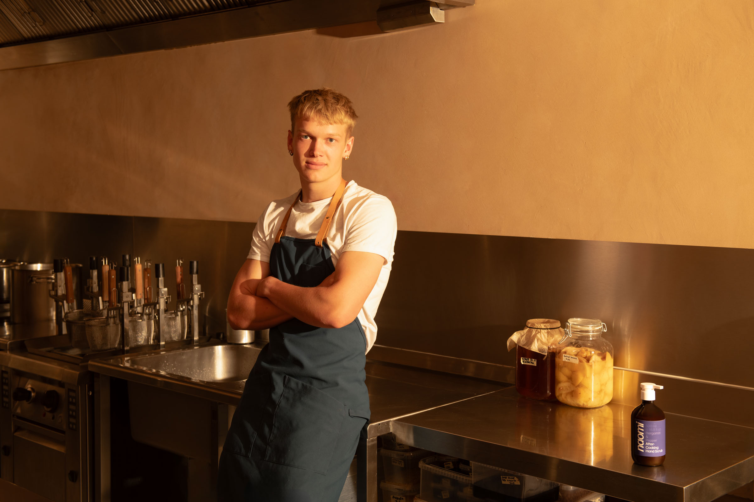 Person in apron standing in a kitchen with jars and utensils.