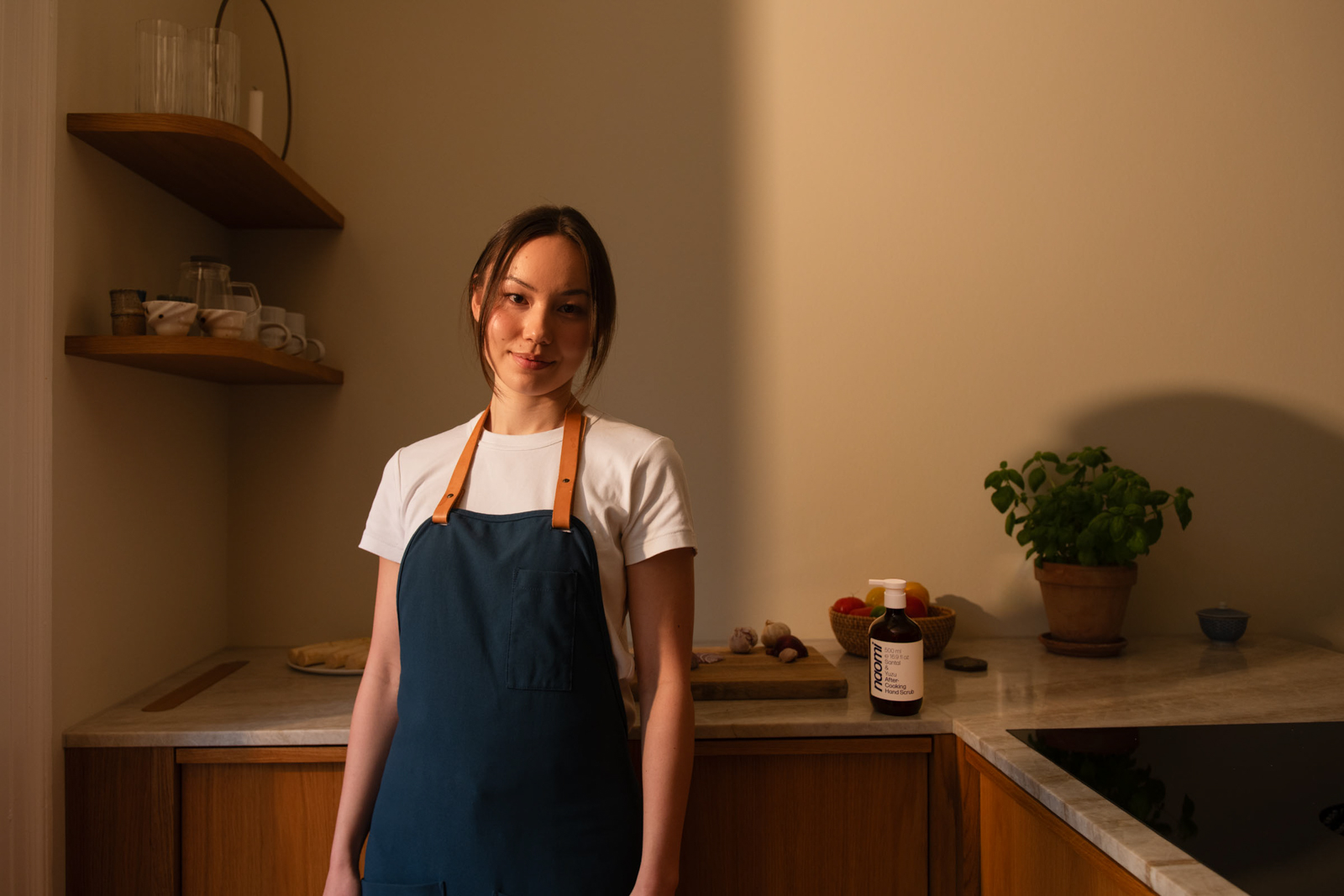 Person in apron standing in a kitchen with shelves and plants.