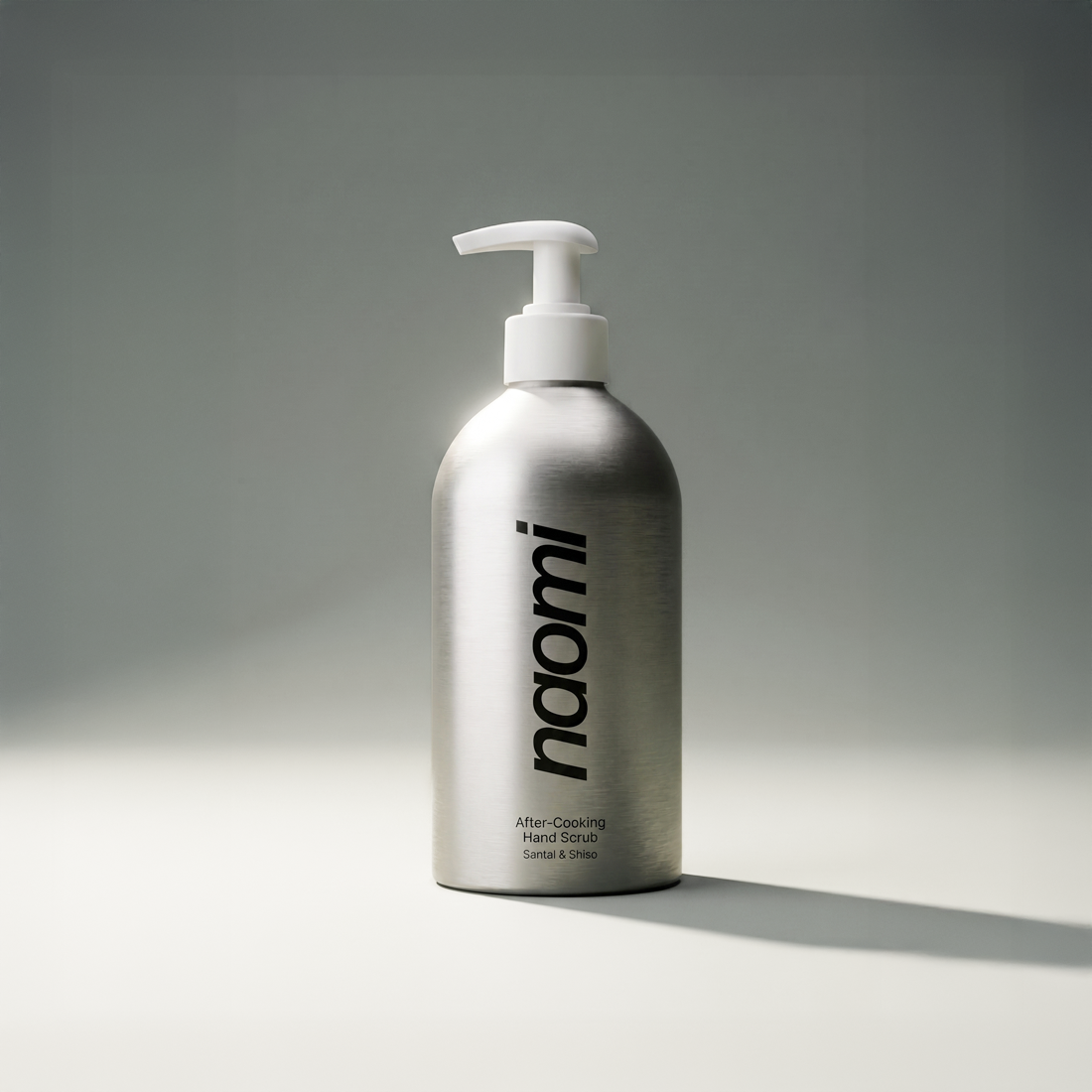 A brushed aluminum pump bottle of Naomi After-Cooking Hand Scrub, Santal & Shiso, against a plain background.
