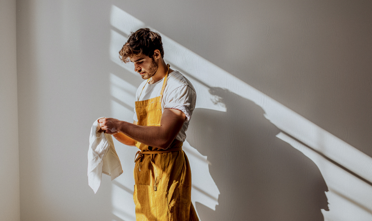 Man in yellow apron holding a cloth, standing in sunlight.