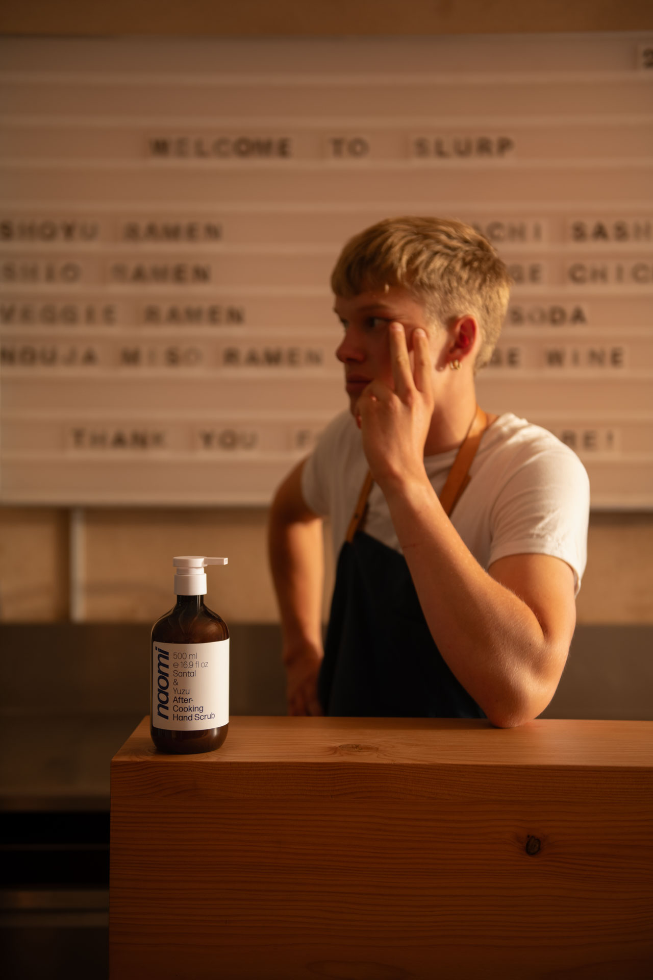 Person in apron standing by a counter with a bottle of hand scrub.