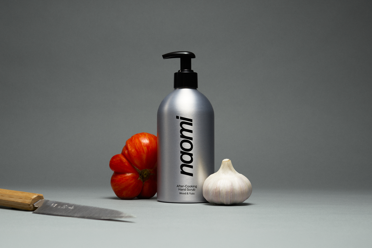 A still life of a silver pump bottle next to a tomato, garlic, and a knife.