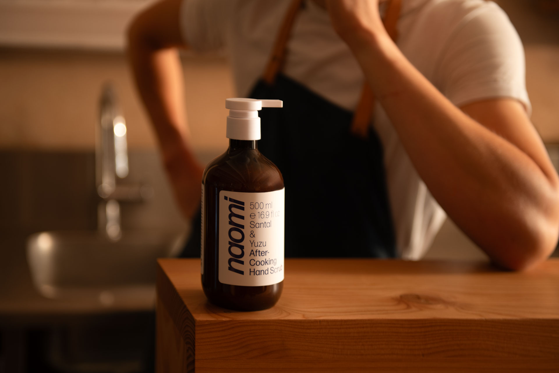 Bottle of hand scrub on a wooden counter with a person in the background.