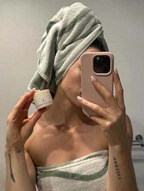 Person with towel on head taking mirror selfie, holding a small container.