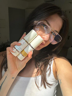 Person holding skincare products, wearing glasses and white top.