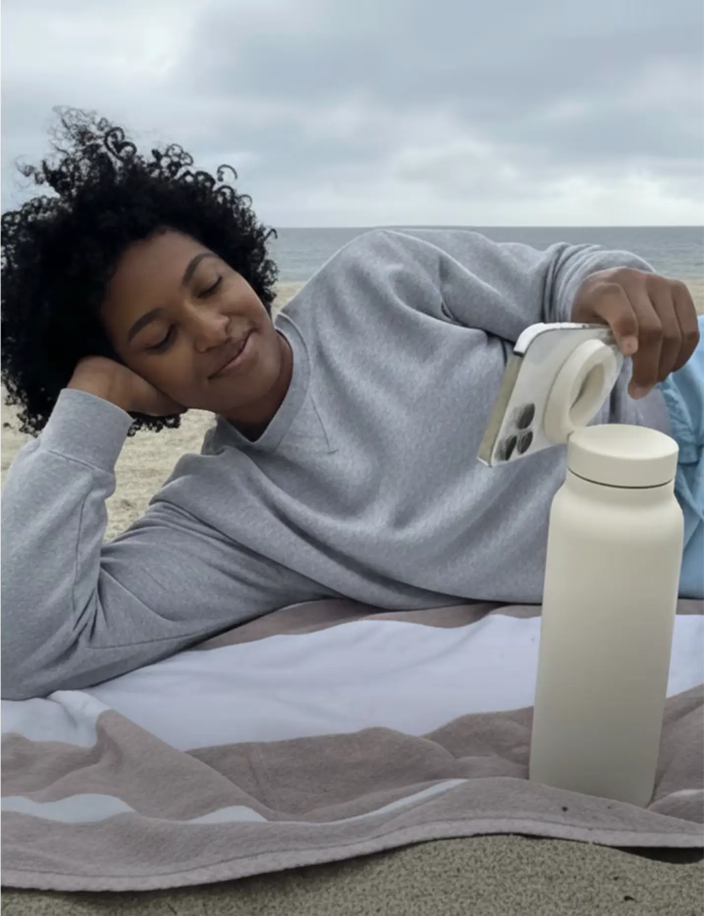 Person lying on a beach towel, taking a photo with a smartphone, near a tall bottle.