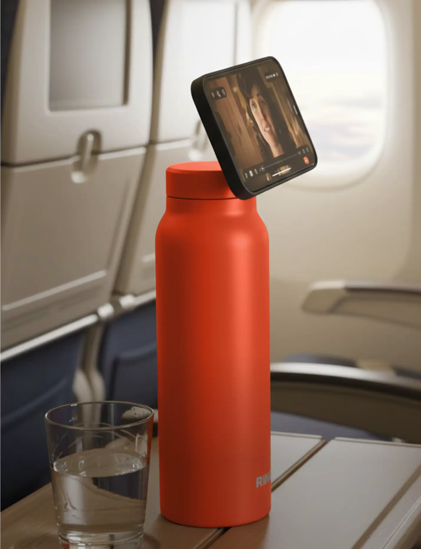Red bottle with phone attached, glass of water on airplane tray table.