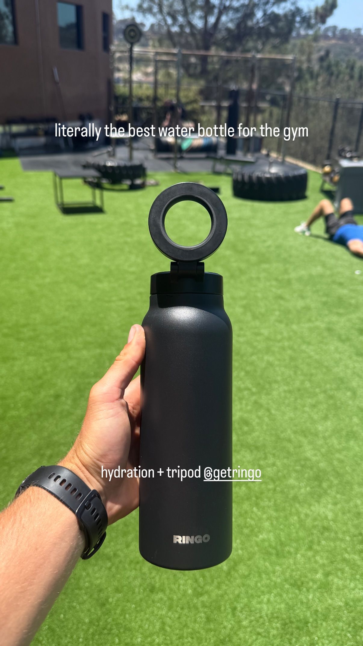 Hand holding a black water bottle with a unique ring handle, gym equipment in the background.