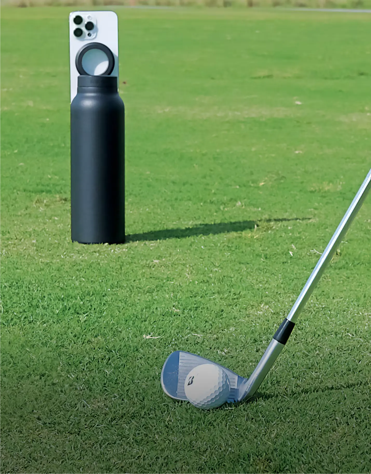 Golf club striking a ball next to a bottle with a mobile phone attached.