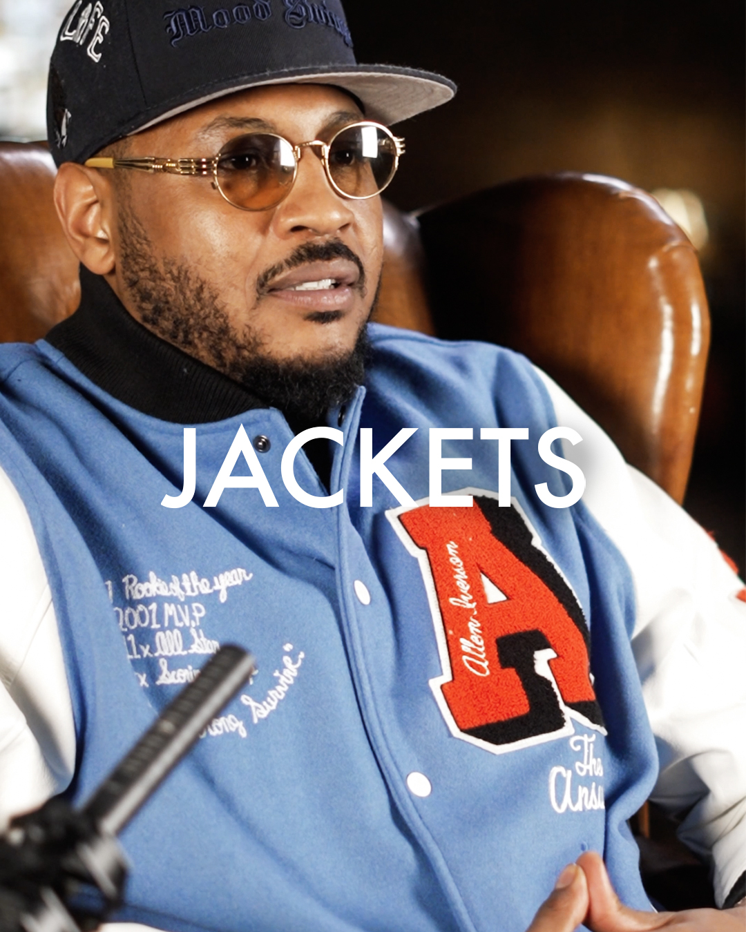 Man in blue varsity jacket with cap and sunglasses, seated in a chair.