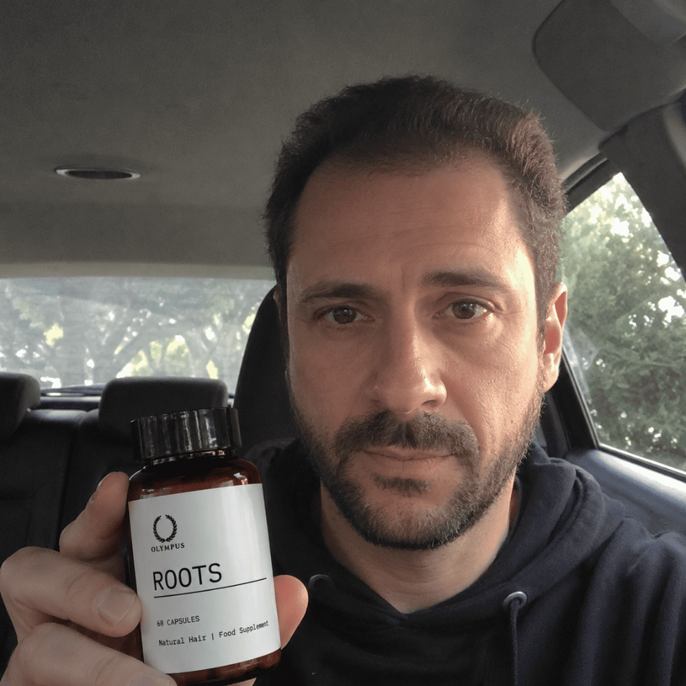 A man in a car holds up a bottle of Olympus Roots Natural Hair Food Supplement.