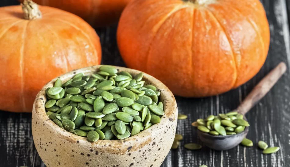 A bowl of green pumpkin seeds sits on a dark wooden surface with whole pumpkins in the background.