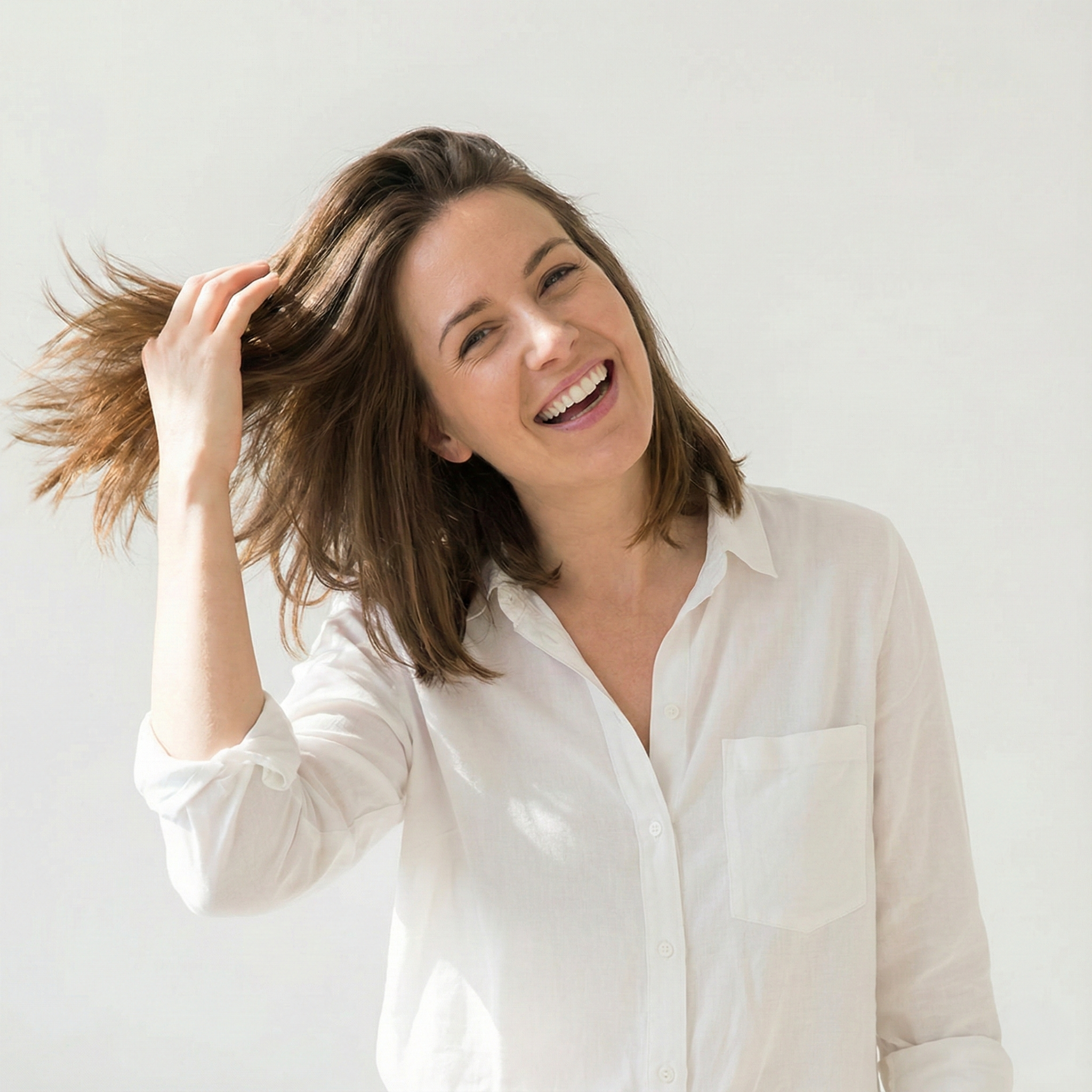 Smiling person in a white shirt, touching their hair, against a plain background.