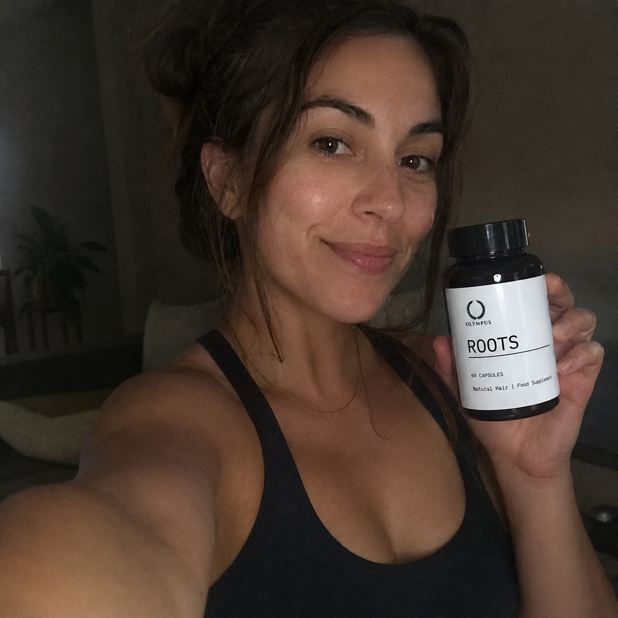 A smiling woman with her hair in a bun holds up a bottle of Olympus Roots hair supplement.