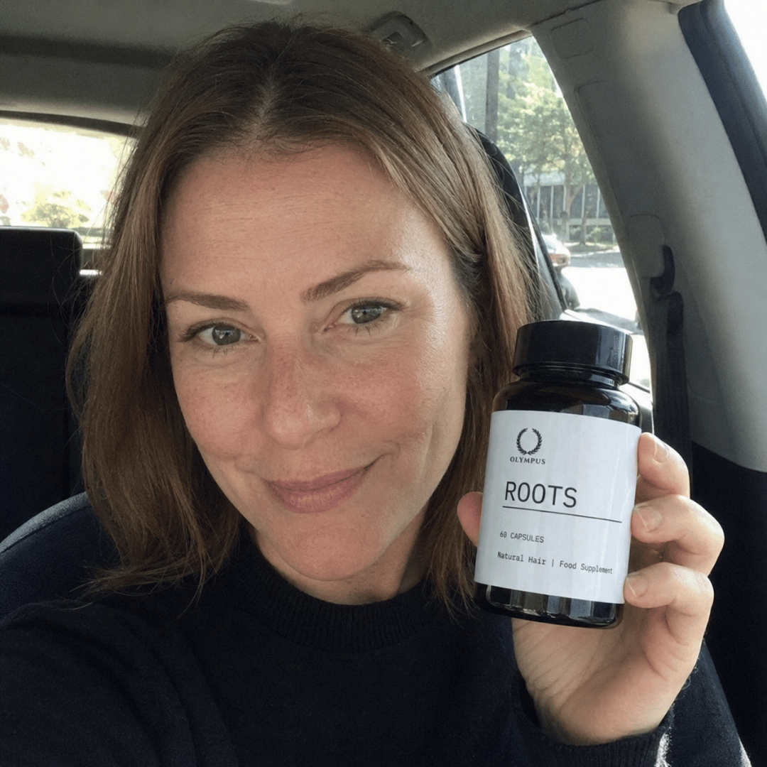 A woman with light brown hair in a car holds up a bottle of Olympus Roots hair supplement.
