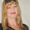 A headshot of a smiling woman with long, blonde hair and bangs, wearing red lipstick.